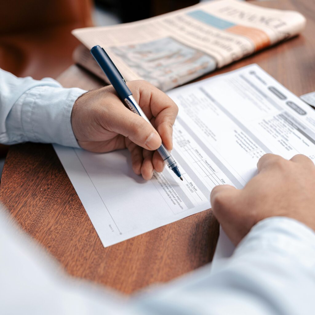 pexels-photo-16282318-16282318 Person reviewing and writing on financial documents with a pen on a wooden desk.